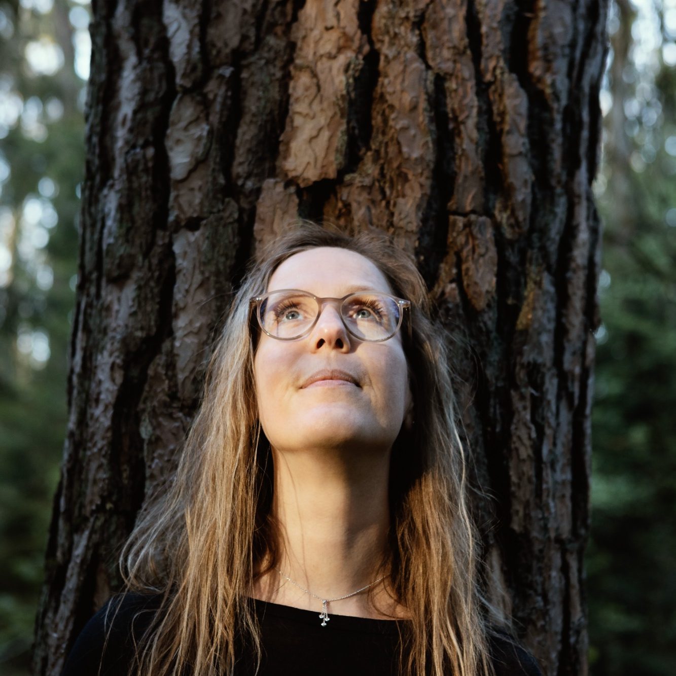 Frau mit Brille schaut nach oben, steht vor einem Baum im Wald.