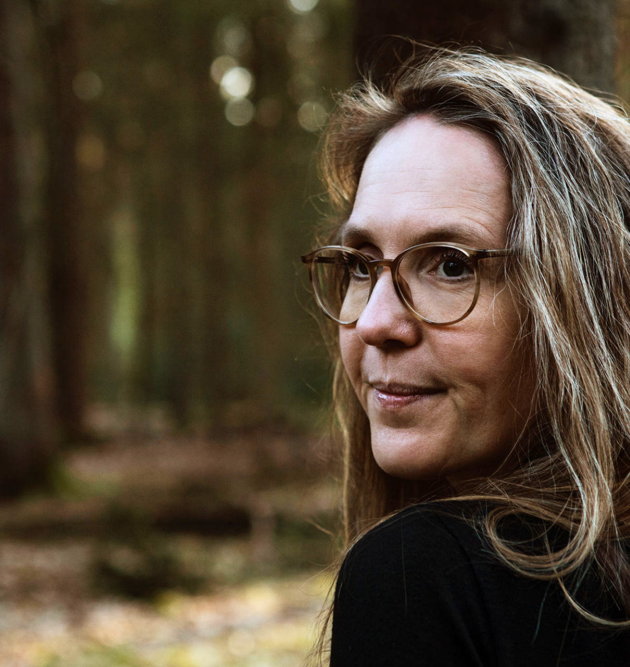 Frau mit Brille, die in einem sonnigen Wald mit Bäumen steht.